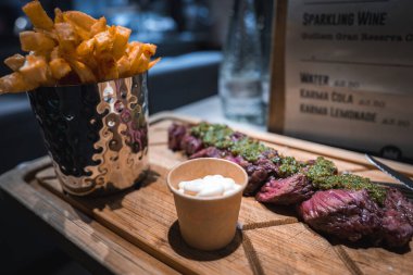 Tahta bir tahtada dilimlenmiş biftek, yeşil chimichurri ve kremalı sos var. Metal bir fincanda çıtır patatesle birlikte. Arka planda bir menü tahtası görünür.