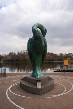 Kuğu, rıhtım, sonbahar ağaçları ve Londra parkında bulutlu bir gökyüzü olan sakin bir gölün yanında dairesel bir kaide üzerinde yeşil bronz bir kuş heykeli..