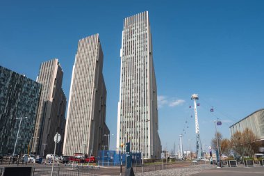 Londra 'da dikey cepheli uzun gökdelenler, Emirates Hava Hattı teleferikleri, asfaltlı bir cadde ve açık mavi gökyüzünün yer aldığı şehir manzarası..
