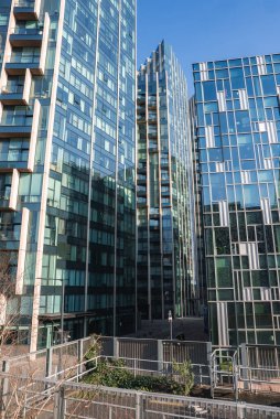 Londra 'da yansıtıcı cepheleri, geometrik tasarımları ve ön planda yeşilliği olan küçük bir çitle çevrili yüksek binalar inşa edildi..