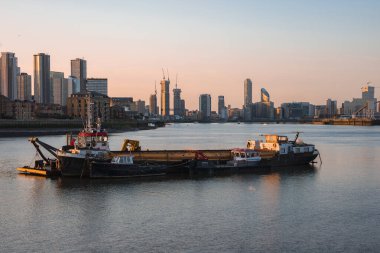 Londra 'da gün batımında Thames Nehri manzarası, demirlemiş tekneler, bir mavna ve sıcak ışığı yansıtan Canary Wharf' ın modern gökdelenleri..