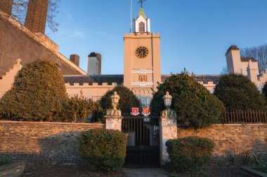 Londra 'daki Royal Hospital Chelsea' de merkezi bir saat kulesi, altın saat yüzü, süslü demir kapı, budanmış çitler ve mavi gökyüzünün altında bir krema cephesi bulunuyor..