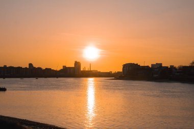 Londra 'daki Thames Nehri' nin üzerinde sakin bir günbatımı. Suda altın yansımalar, modern binalar ve ufuk çizgisinde görünen Çömlek..