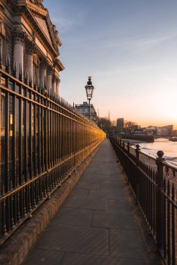 Günbatımında Londra 'da bir nehir kenarı geçidi, tarihi bir bina, geleneksel bir sokak lambası ve modern binaların ötesinde Thames nehri var..
