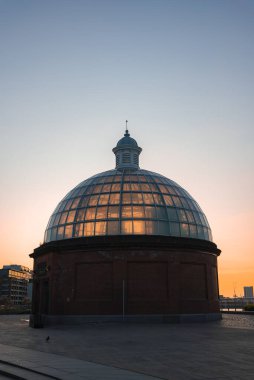 Greenwich Ayak Tüneli 'nin kubbeli cam ve tuğla girişi günbatımı renkleriyle aydınlatılıyor. Arka planda modern binalar ve Thames Nehri görülebilir..