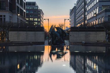 Londra 'da günbatımı manzarası, yansıtıcı su özelliği, soyut heykel, çağdaş ofis binaları ve sokak lambaları içerir..