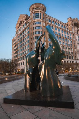 Soyut biçimlere sahip modern bir bronz heykel Londra plazasında duruyor. Arkasında, kıvrımlı mimarisi ve ağaçları olan büyük bir bina sahneyi tamamlıyor..