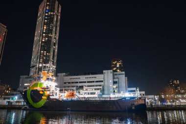 Gece Londra 'da sakin sulara yanaşan parlak ışıklı bir gemi, uzun bir gökdelen ve suya ışık yansıtan geometrik beyaz bir bina..