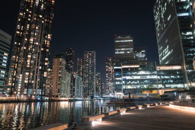 Aydınlatılmış pencereli modern gökdelenler Londra, Canary Wharf 'taki sakin rıhtım sularını yansıtıyor. Yanmış ahşap bir tahta yol suyun kenarını çiziyor..