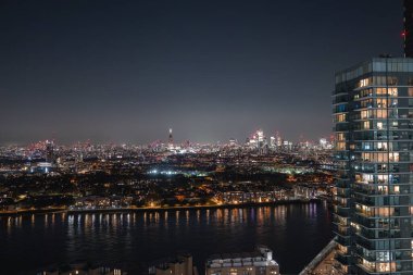 Gece aydınlanan Londra silueti Parça, parlayan yüksek binalar ve Thames nehri ön plandaki şehir ışıklarını yansıtıyor..
