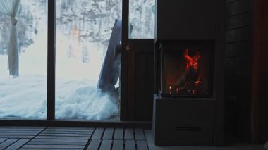 Modern bir şömine, sıcak bir odanın içinde sıcak bir parıltı yayar. Dışarıdaki zemin karla kaplı, büyük cam kapılardan görülebiliyor, titreyen alevler hareket katıyor..