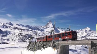 Kırmızı bir Gornergrat treni Zermatt, İsviçre 'de yüksek bir demiryolu üzerinde seyahat eder, arka planda Matterhorn, kar kaplı yamaçlar ve açık gökyüzü ile çevrilidir..