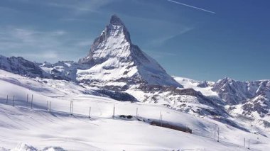 Kızıl bir Gornergrat treni, İsviçre Alpleri 'ndeki Matterhorn' un tabanında, açık mavi gökyüzü altında karla kaplı manzaralarla dolu bir demiryolu boyunca seyahat eder..