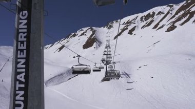 Bir kayak asansörü Alplerdeki karlı bir dağ yamacında yukarı doğru hareket eder ve kapalı gondollar yolcu taşır. Engebeli arazi ve mavi gökyüzü görünür..