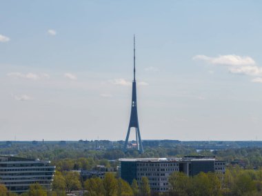 Riga Radyo ve TV Kulesi, modern binalar ve yemyeşil yemyeşil, arka planda dağınık bulutlarla çevrili açık mavi gökyüzüne karşı yükseliyor..