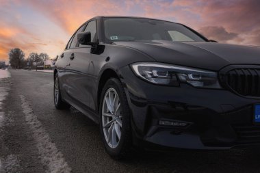 Üzerinde keskin LED farlar, böbrek ızgarası, gümüş alaşımlı tekerlekler ve mavi bir plaka bulunan asfalt bir yolda park edilmiş siyah BMW G20 320 model. Nadir ağaçlar ve uzak binalar bulutlu bir gökyüzünün altında, kısmen karla kaplı yol kenarı sularıyla görülebilir.