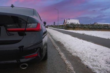 Siyah bir BMW G20 320 'nin arka görüntüsü görünür stop lambaları ve egzoz borusu, karla kaplı çatıları ve çitleri olan evlerin yakınındaki karlı bir yolda park edilmiş..