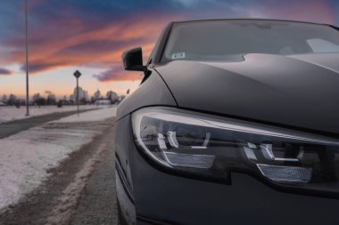 Siyah bir BMW G20 320 'nin ön sağ görüşü. Karlı bir yolda park edilmiş. Özellikler arasında parlak farlar, bulutlu bir gökyüzü, seyrek ağaçlar ve sokak ışıkları yer alıyor.