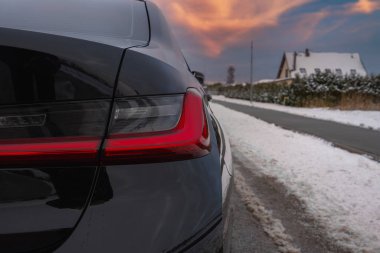 Siyah bir BMW G20 'nin arka sol arka lambasının yakın görüntüsü. Kar kaplı bir yolda park edilmiş, kışın banliyöde bir evi ve çitleri var..