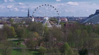 Riga Zafer Parkı, kiraz çiçeği mevsiminde, bir dönme dolap, St. Peter Kilisesi kulesi ve arka planda Letonya Ulusal Kütüphanesi yer alır..