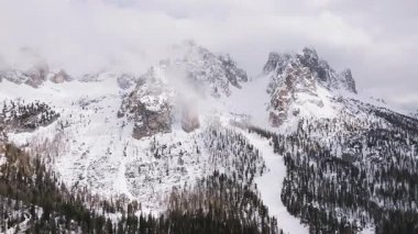 Cortina yakınlarındaki sivri Dolomite tepelerinin havadan görünüşü kısmen bulutlarla örtülü, karlı yamaçlarda daima yeşil ormanlar ve yumuşak kamera hareketleri var..