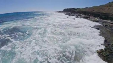 Dinamik dalgalar, Oahu ya da Hawaii 'deki Kauai' de açık mavi gökyüzünün altında seyrek yeşilliğe sahip kayalık bir tepenin yakınındaki engebeli volkanik kayalara çarpar..