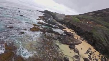 Enerji dalgaları Hawaii 'nin kıyı şeridi boyunca uzanan siyah volkanik kayalara çarpar. Yeşil kayalıklar ve bulutlu bir gökyüzü dramatik bir manzara yaratır..