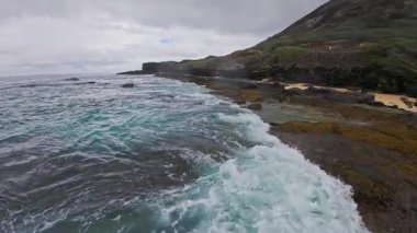 Dinamik okyanus dalgaları, Oahu ya da Kauai 'deki sarp, yeşil bir tepenin yakınındaki karanlık volkanik kayalara çarpar..