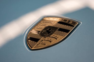 Porsche ambleminin açık mavi bir yüzeydeki yakın görüntüsü, altın ve siyah tasarımını, metalik dokusunu ve cilalı kaplamasını sergiliyor..