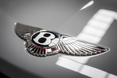 Bentley logosunun detaylı görüntüsü kalın B, gümüş kanatlar ve siyah oval, parlak gri bir araba yüzeyi üzerindeki pencere ızgarasını yansıtıyor..