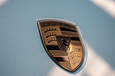Açık mavi bir yüzeyde bulunan Porsche ambleminin detaylı bir görüntüsü, kırmızı ve siyah aksanlı ve metalik desenli altın ve siyah tasarımını gözler önüne seriyor..
