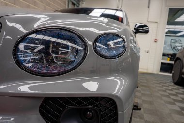Kapalı bir galeride gri Bentley Bentayga 'nın yakın görüntüsü, çift dairesel farları, cilalı yüzeyi ve fayanslı döşemeleri vurguluyor..