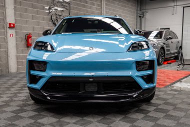 Açık mavi bir Porsche Macan kapalı bir yere park etmiş gri tuğlalı bir duvar, aslan amblemi ve arka planda yangın söndürücü var..