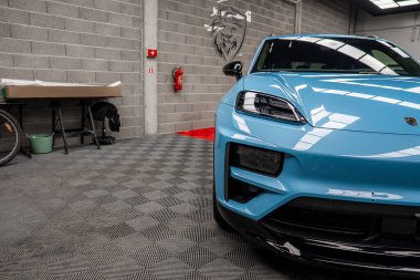 Beton duvarları, aslan amblemi ve görünür arkaplan ögeleri olan modern bir sergi salonunun kareli zeminine park etmiş mavi bir Porsche Macan 'ın ön görüntüsü..