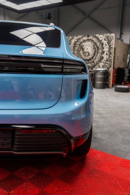 Açık mavi bir Porsche Macan 'ın kırmızı bir zemine park edilmiş arka görüntüsü. Arkaplanda desenli bir duvar, lastikler, araçlar ve tepeden aydınlatma yer almaktadır.