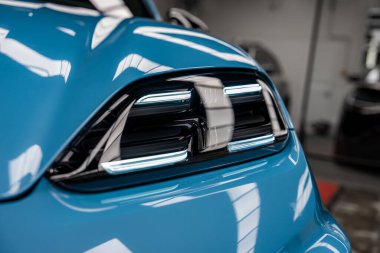 Kapalı mekanda bir Porsche Macan süper arabasının ön stop lambası, keskin LED elementler, parlak siyah konutlar, ve tepedeki ışıkların yansımaları.