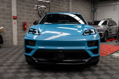 Canlı mavi bir Porsche Macan, arka planda gri tuğlalı duvarlar, damalı zemin ve gümüş renkli Bentley Bentayga ile kapalı bir galeride sergileniyordu..