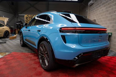 Mavi bir Porsche Macan kırmızı kareli bir paspasın üzerine park etmiş, arka lamba barını gösteriyor. Arka planda bir bej araba ve otomotiv ekipmanı görünür.