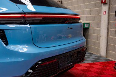 Mavi Porsche Macan Turbo kırmızı ve siyah kareli zemine park etmiş. Arka planda yangın söndürücü ve yeşil kutu olan beton duvar..