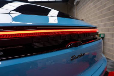Kırmızı LED stop lambalı mavi bir Porsche Macan Turbo, Porsche ve Turbo rozetli, tuğla duvarlı ve döşemeli bir sergi salonunda.