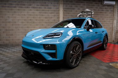 Pürüzsüz tasarım elementleri olan parlak mavi bir Porsche Macan kapalı bir yerde kareli zeminde oturuyor. Tepedeki ışıklandırma onun parlak yüzeyi ve çatı aksesuarını vurgular.