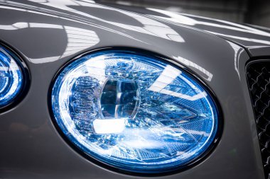 Bentley Bentayga 'nın yansıtıcı elementler ve kapalı bir alanda parlak bir ızgarayla mavi ışık saçan farının detaylı görüntüsü..