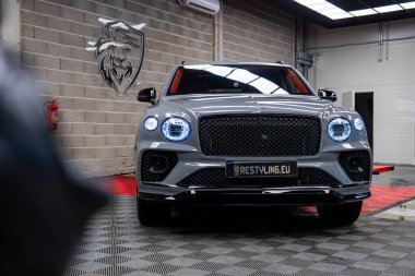 İyi aydınlatılmış kapalı bir garajda kareli zeminde gümüş renkli Bentayga lüks bir SUV. Tuğla duvarda kırmızı bir platform ve aslan amblemi görülebilir..
