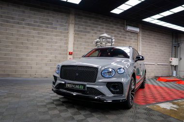 Açık gri bir Bentley Bentayga SUV, parlak siyah ızgara ve çatı rafı, evin içinde sergilenen tuğla bir duvar ve arka planda kareli zemin paspası.