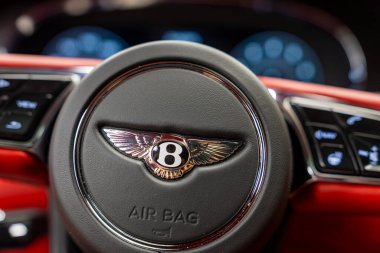Kırmızı iç mekan, kontrol düğmeleri ve bulanık dijital gösterge paneli ile çevrili ikonik B logosunun bulunduğu Bentley direksiyonun yakın görüntüsü.