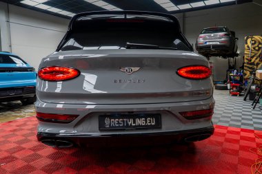 Bentayga 'nın gri Bentayga' sı kırmızı kareli paspasın üzerine park etmiş ve Bentley logosu görünüyor. Mavi Porsche Macan ve araba kaldıracı temiz atölyede görünür..