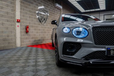 Galeride mavi farları olan gri bir Bentley Bentayga SUV. Özellikler arasında tuğla duvar, aslan amblemi, kırmızı halı ve şebeke tavan ışıklandırması yer alıyor..