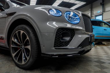 Aydınlatılmış mavi farlı gri bir Bentley Bentayga arka planda mavi bir Porsche Macan ile kapalı bir yere park edilmiştir..
