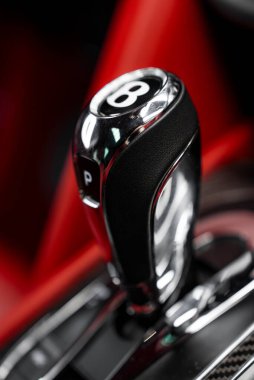 Bentley vites değiştiricisinin krom ve siyah deri içinde, ikonik B logosunun olduğu yakın plan görüntüsü. Arka planda kırmızı iç aksanlar görünür..