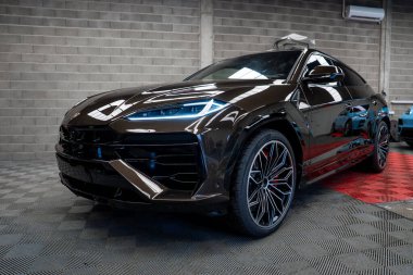 Koyu kahverengi bir Lamborghini Urus, parlak bir kapağı olan kırmızı ve siyah kareli bir zemine park edilmiştir. Gri tuğla bir duvar ve havalandırma sistemi görülebilir..